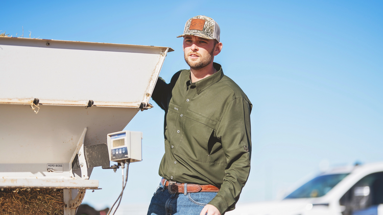 Man in a green long sleeve shirt and camo hat standing on the back of a truck outside 
