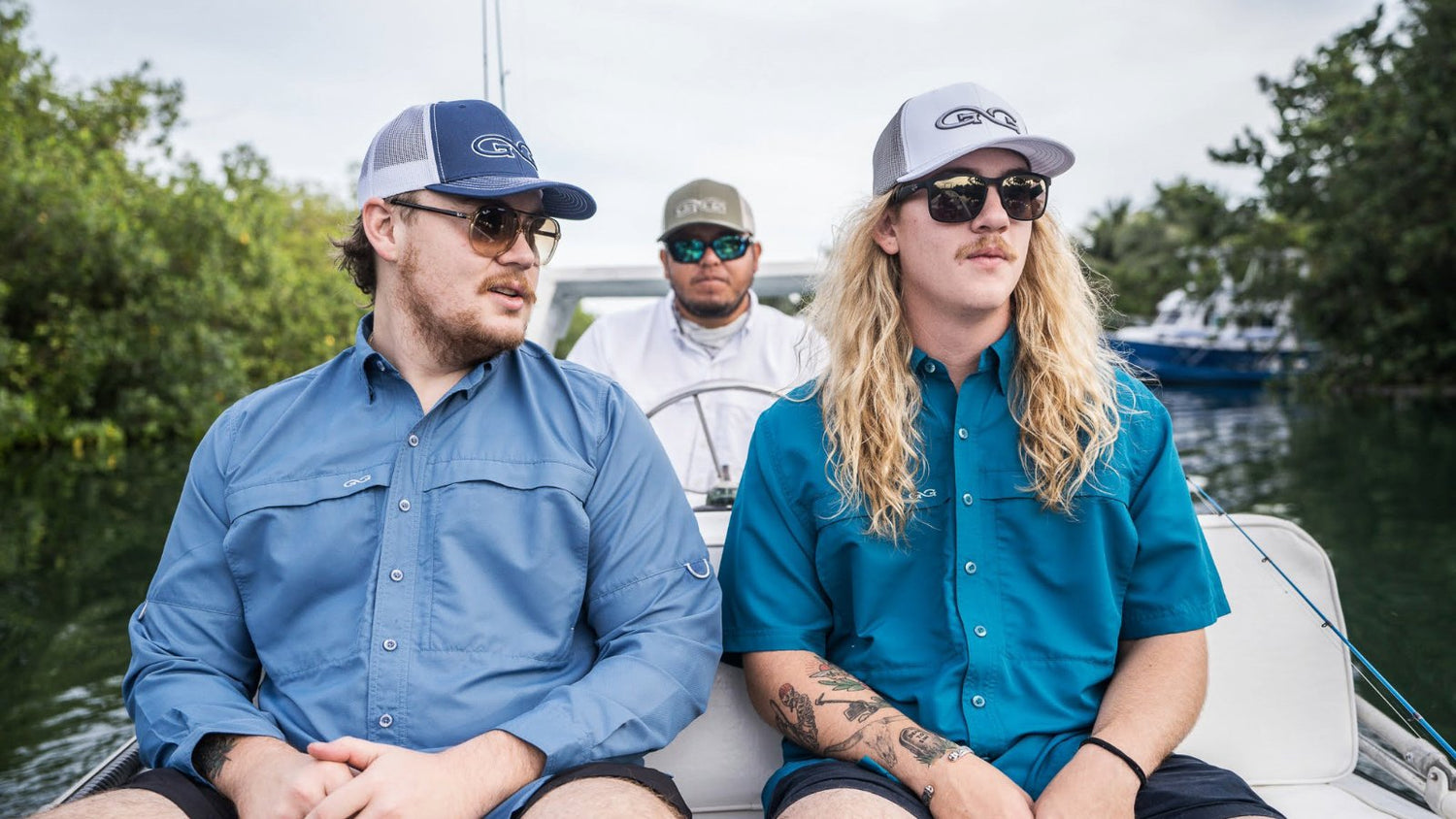 “ALL MICROFIBER SHIRTS.” Two men sit on the bow of a boat wearing GameGuard MicroFiber Shirts and GG meshback caps. A third man is steering in the background. Trees line the waterway, and fishing rods are visible.