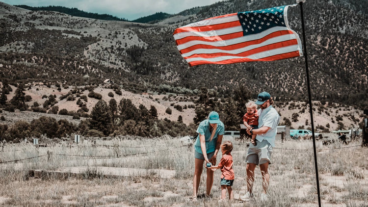 AMERICA COLLECTION. Family stands under US flag in GameGuard MeshBack Caps, performance outdoor shirts, and Travel Shorts. Patriotic outdoor apparel, red white & blue gear, GameGuard USA collection, UPF shirts, moisture-wicking, summer adventure clothing.