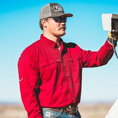 Man wearing a red long-sleeve work shirt, jeans, and cap operating ranch equipment under a clear blue sky.