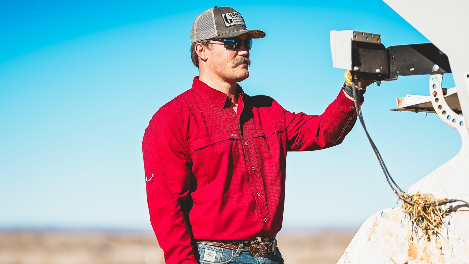 A man wearing a red GameGuard Maverick Shirt, jeans, gloves, sunglasses, and a cap works outdoors, adjusting ranch equipment under a bright blue sky.