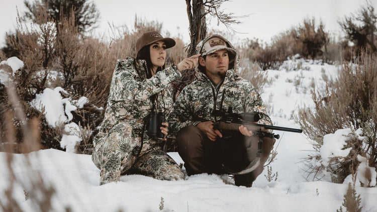 Man and women in the cold snow wearing GameGuard Guia Grande Jackets, Pants, and GameGuard Caps staying warm in the outdoor cold environment while hunting. 