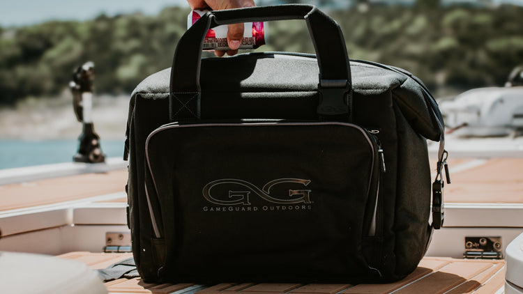 FIELD GEAR & COOLER BAGS. Two paddleboards rest on a wooded shoreline with paddles and GameGuard cooler bags on top, surrounded by dense green foliage and calm water.