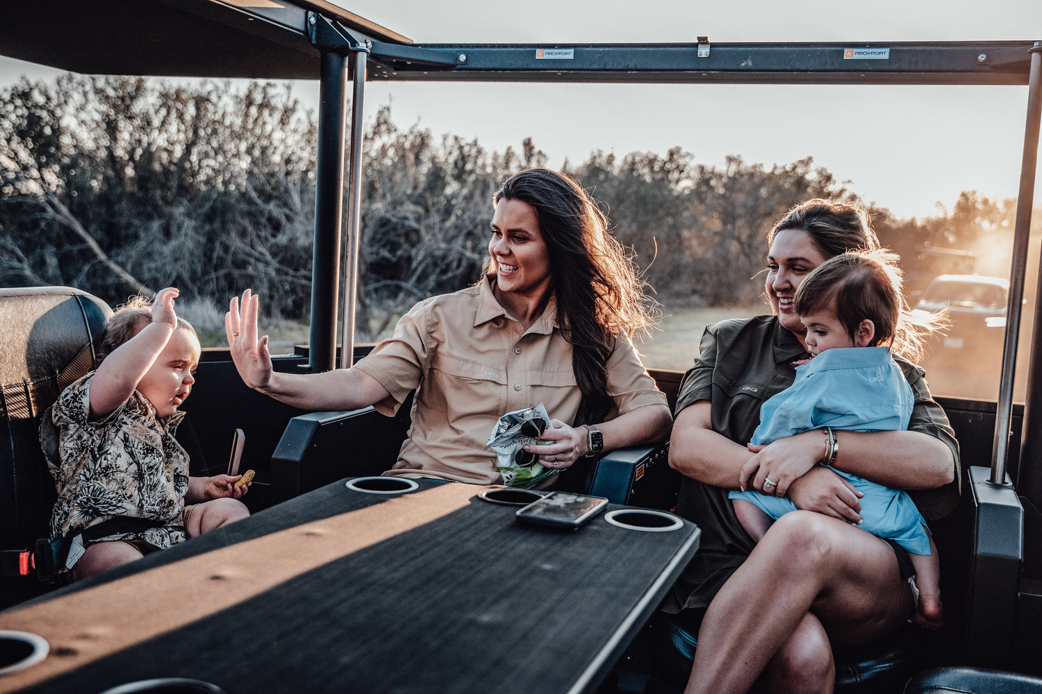 Two women and two young children ride in an open utility vehicle at sunset. One woman wears a khaki GameGuard shirt, the other an Agave GameGuard shirt, laughing and smiling.