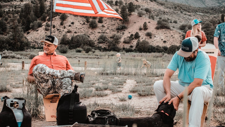 ALL PRODUCTS. Two men relax outdoors wearing GameGuard Original Shirt and MeshBack Caps. GameGuard Digital camo pants and hunting gear shown. Dog nearby, kids play behind. Outdoor apparel, camo clothing, lifestyle caps, and field-tested gear.