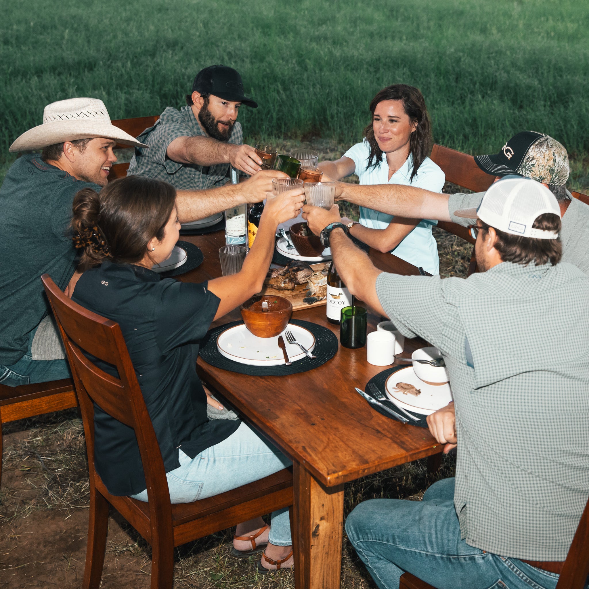 Group of people sitting around a wooden table raising a glass outside