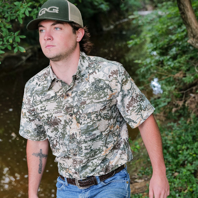 Cargue la imagen en el visor de la galería, an wearing a GameGuard Digital Explorer Shirt for men, standing outdoors near a stream, with greenery in the background. The shirt features a camouflage pattern.
