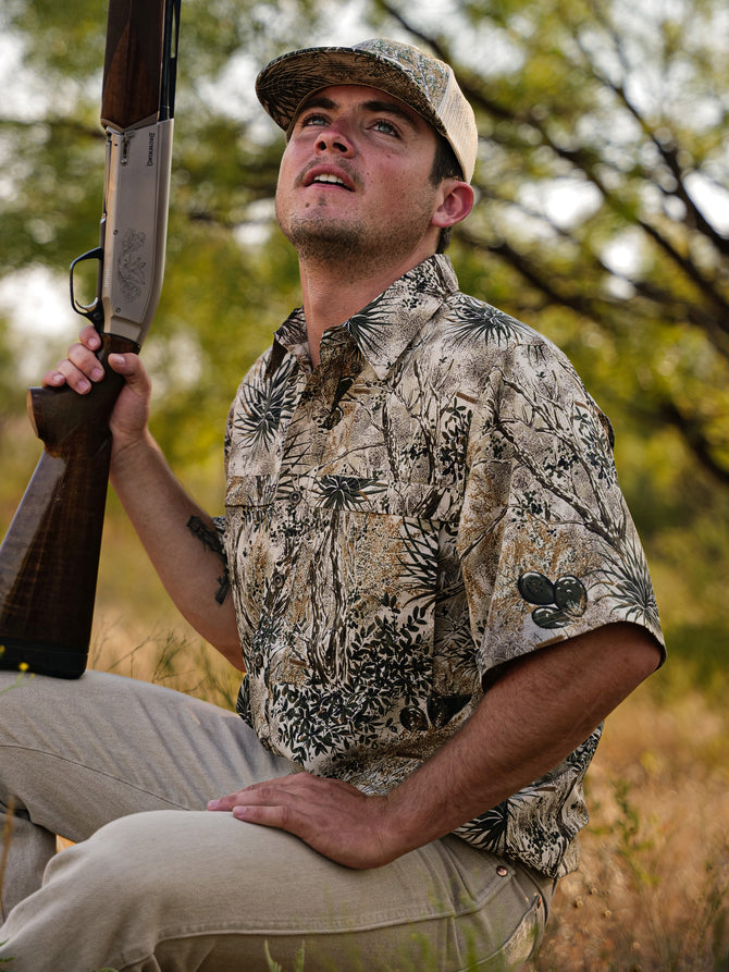 man sitting in brush wearing GameGuard cap and GameGuard MicroFiber Shirt holding shotgun